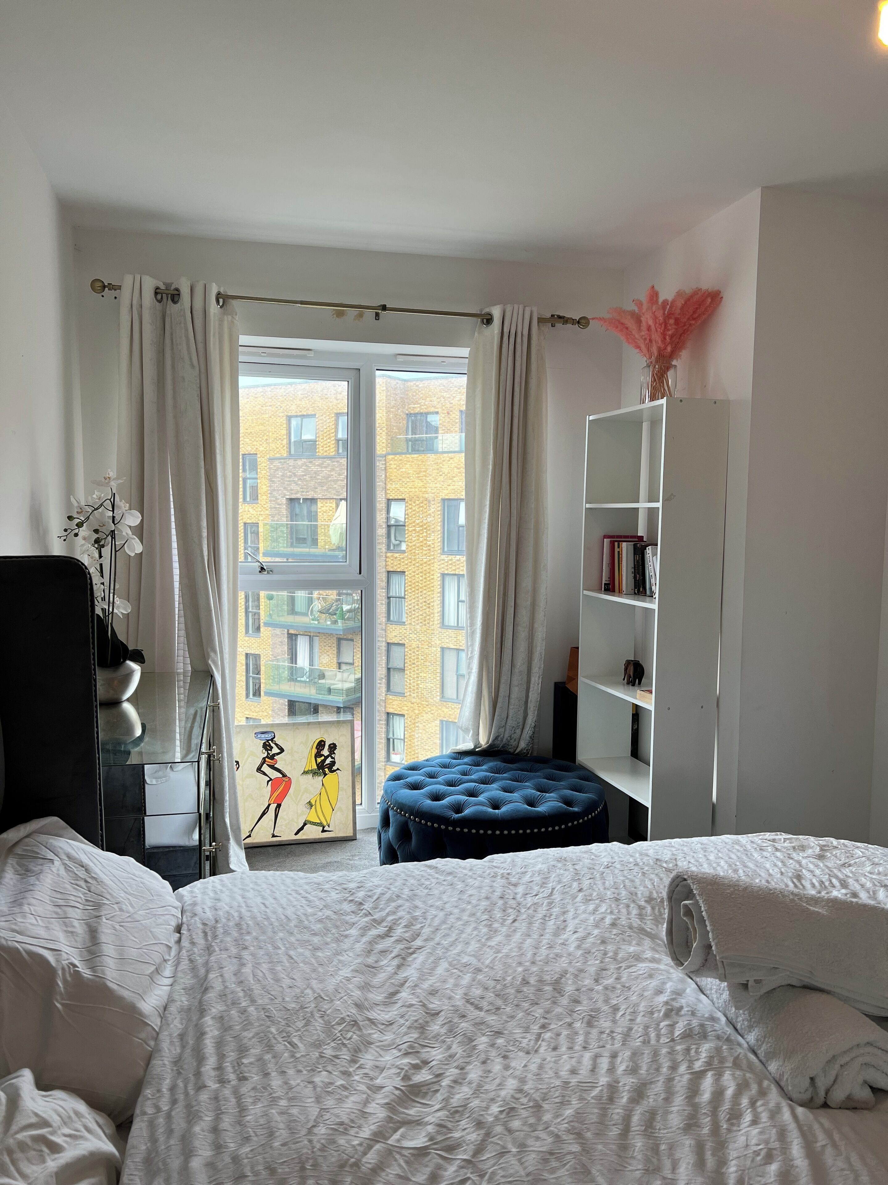 A hotel room with a large window, a bed with white bedding, a blue ottoman, a white wardrobe, and a small bookshelf.