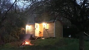 A small, single-story cabin with a visible door and a lit interior, surrounded by trees and a grassy area. - Apple hut, romantic shepherd hut retreat with log burner, hot tub, pet friendly! (Paignton)