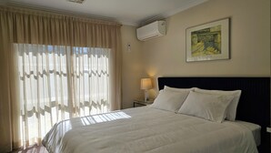 A hotel room with a bed, a nightstand with a lamp, a painting on the wall, and an air conditioning unit. - Modern 2BR/1BA/2+CAR Home in Central Canberra (Campbell)