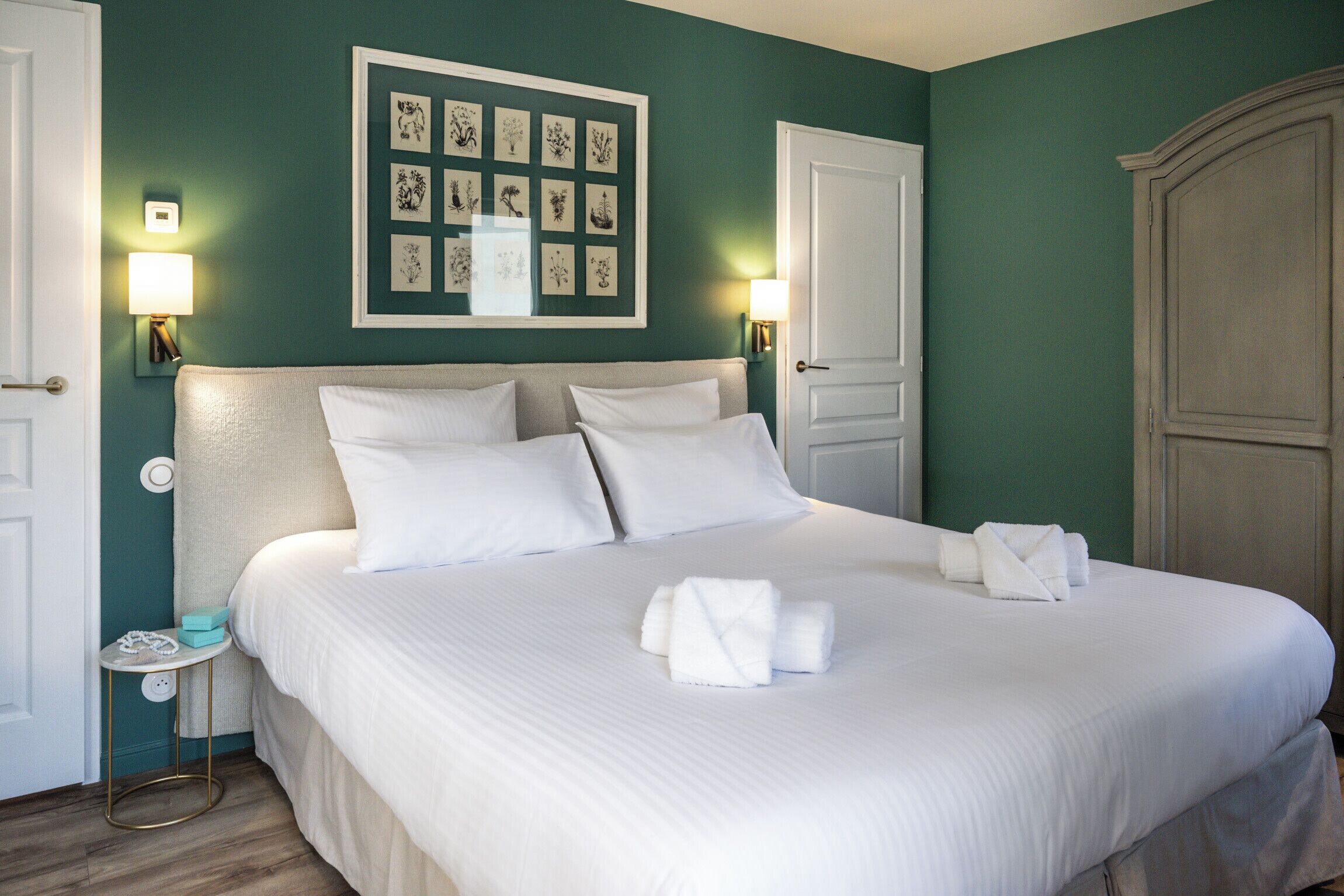 A neatly made bed with white linens and towels, a framed botanical print on the wall, and a wooden wardrobe.