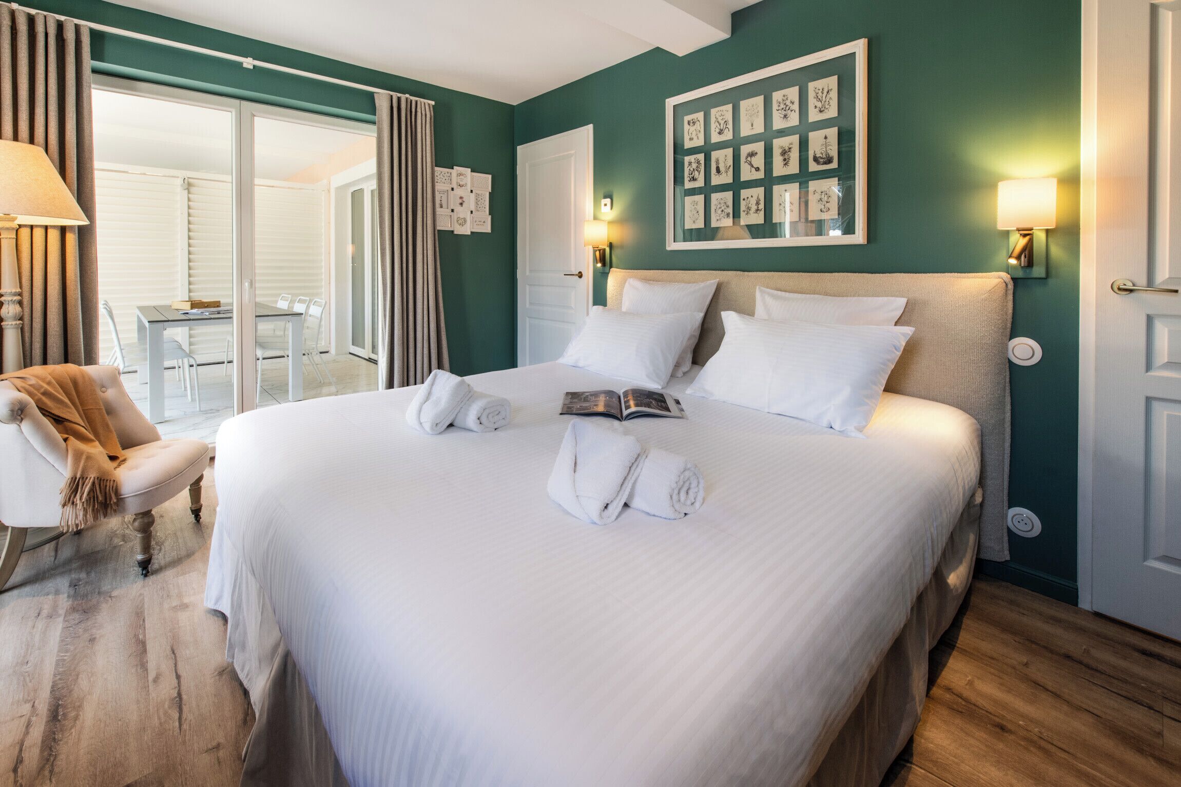 Una cama bien hecha con ropa de cama y toallas blancas, una mesita de noche con un libro y una pared decorada con cuadros.