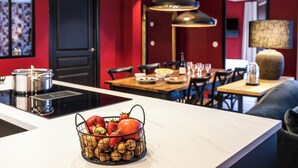 Moderni keittiö, jossa on ruokailutila. Keittiössä on muun muassa keskeinen saareke, jossa on hedelmäkori, musta liesi ja ruokapöytä tuoleineen.