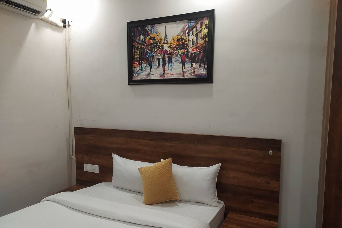 Hotelkamer met een houten hoofdeinde, een bed met wit bedlinnen, één geel kussen, en een lijst met daarin een schilderij van een straatbeeld boven het bed.