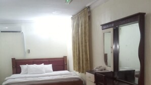 A hotel room with a bed, a desk, a chair, a mirror, and a window with curtains. - Biggie Hotel and Suites  (Lagos)