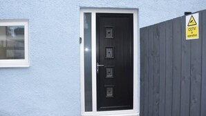 Une porte noire avec un cadre blanc, une fenêtre avec des stores et un panneau d’avertissement indiquant la présence d’un système de vidéosurveillance sur une clôture grise.