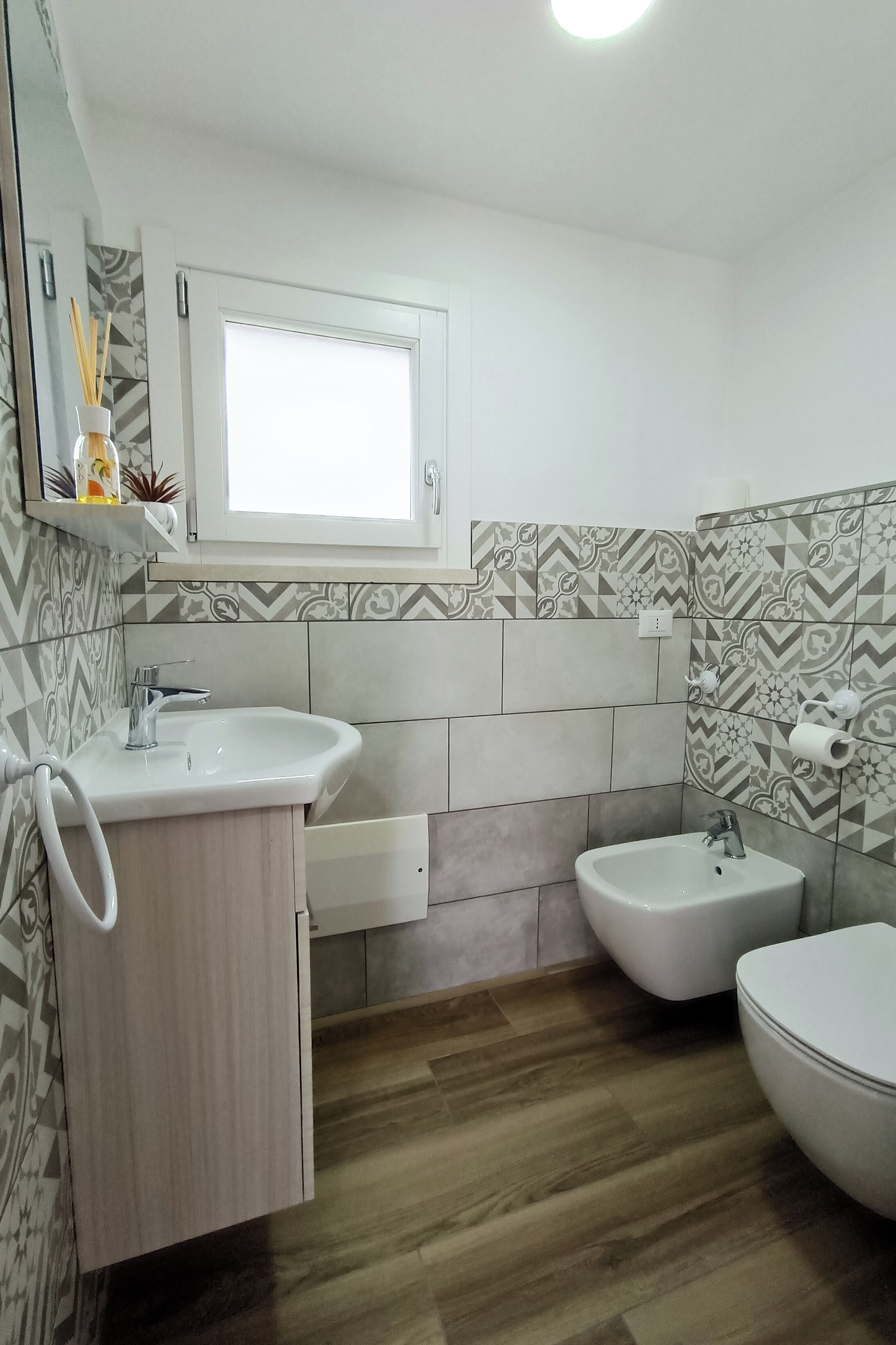 Een badkamer met een witte wastafel, een bidet en een toilet. Er is een raam boven de wastafel en een spiegel aan de muur.