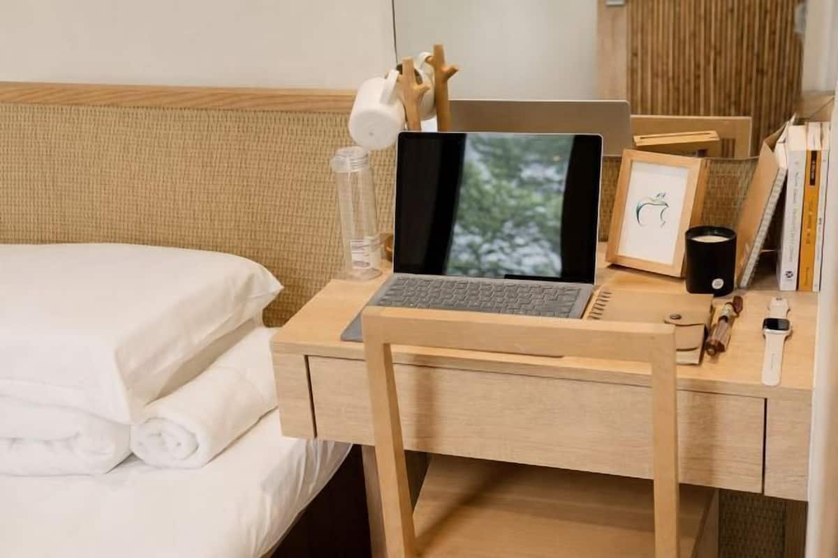 A hotel room with a bed, a wooden desk with a laptop, a smartphone, a picture frame, and a small plant.