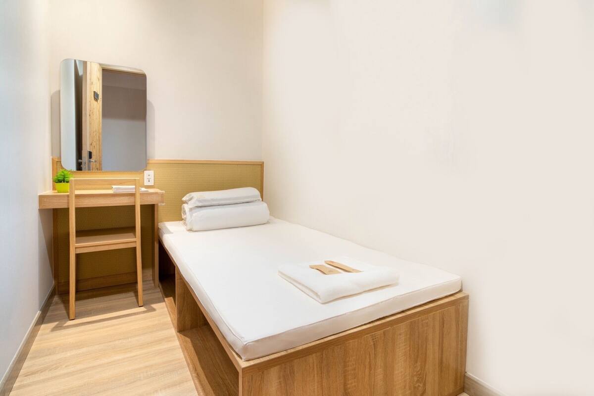 A small, neatly arranged bedroom with a single bed, a wooden nightstand, a mirror, and a small shelf.