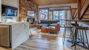 Eine moderne Küche mit Steinwand, großem Fernseher und Kamin. Ein Wohnbereich mit Sofa und Pouffen.