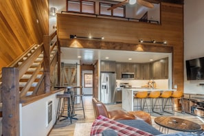 Amplia sala de estar con una escalera de madera, una cocina con electrodomésticos modernos y una zona de bar con taburetes.