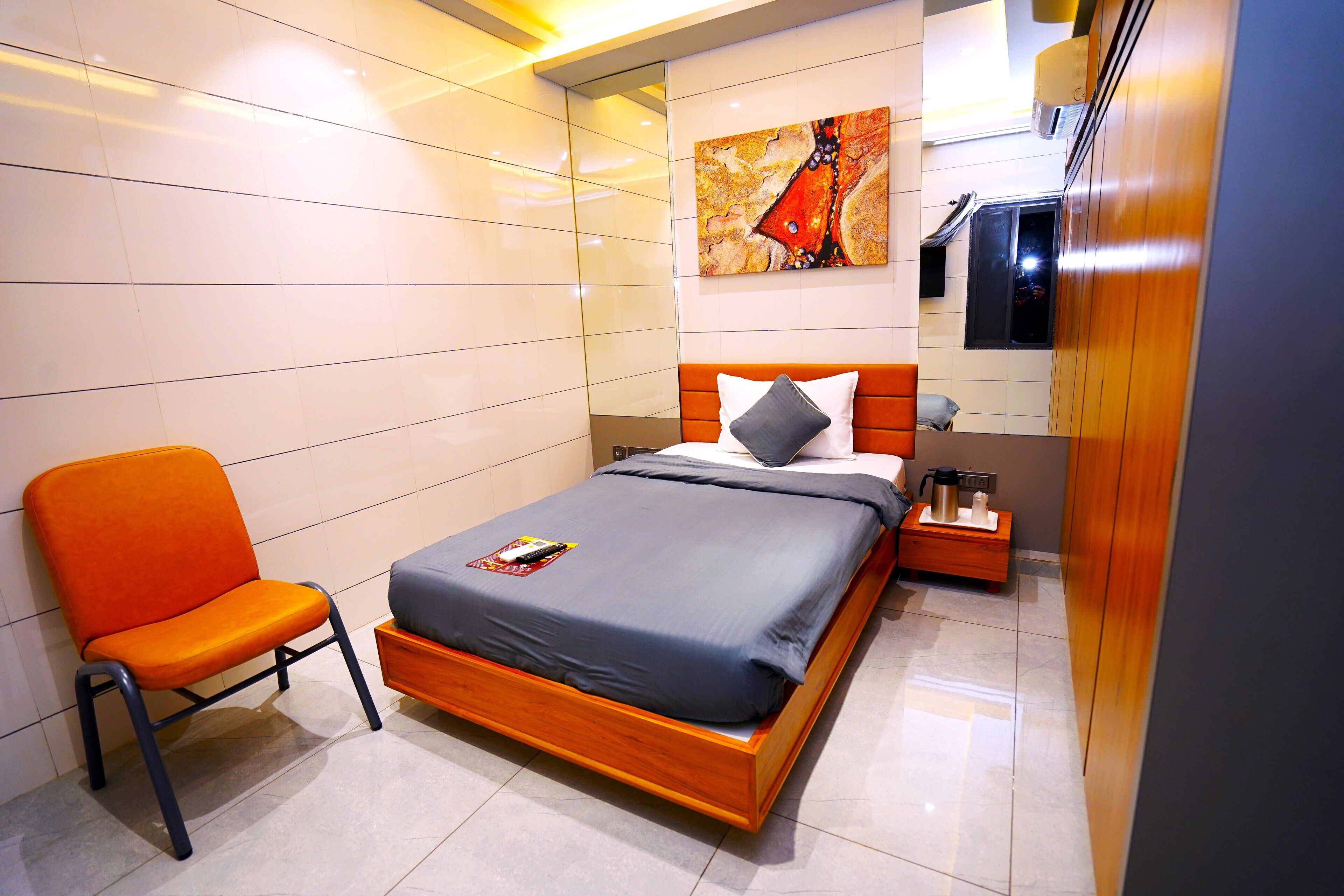Una habitación de hotel moderna con una cama de madera, un colchón gris, una silla naranja, una mesita de noche con una lámpara y un cuadro en la pared.