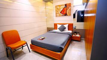Una habitación de hotel moderna con una cama de madera, un colchón gris, una silla naranja, una mesita de noche con una lámpara y un cuadro en la pared.