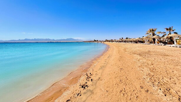 Una playa con agua turquesa cristalina, una orilla de arena y una hilera de palmeras y sombrillas.