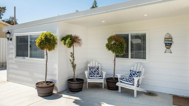 Een modern huis met een overdekte veranda, twee potplanten en twee witte Adirondack stoelen met opvallende kussens.