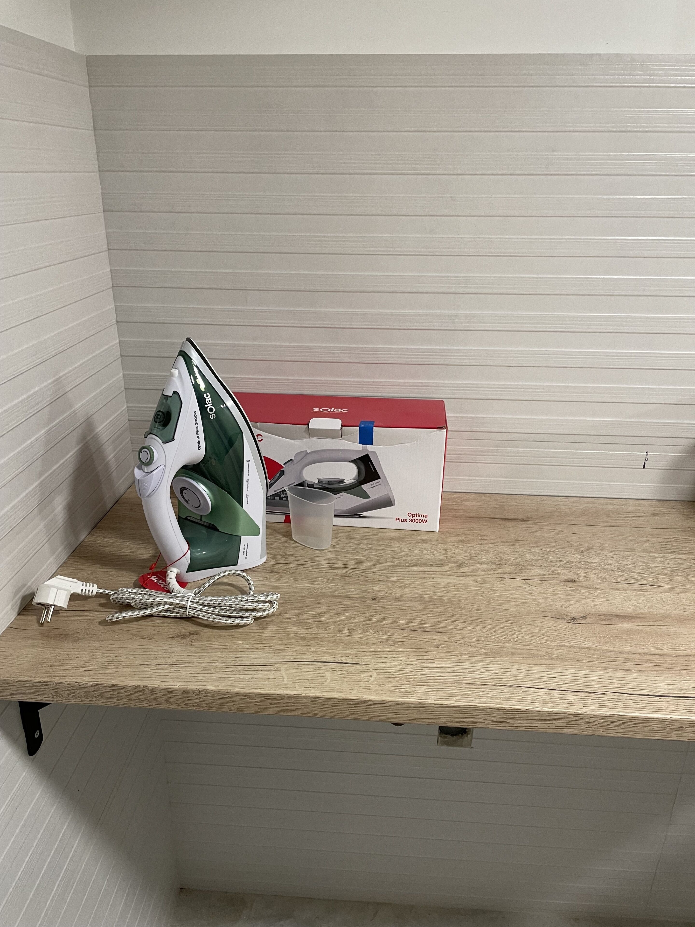 A wooden shelf with an iron, a box, and a power cord in a room with lined walls.