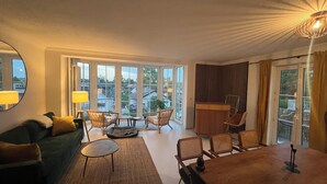 A modern living room with a green sofa, a round coffee table, and a dining table with chairs. Large windows offer a view of the neighborhood. - 2 bedroom apartment in herrsching am ammersee (Herrsching am Ammersee)