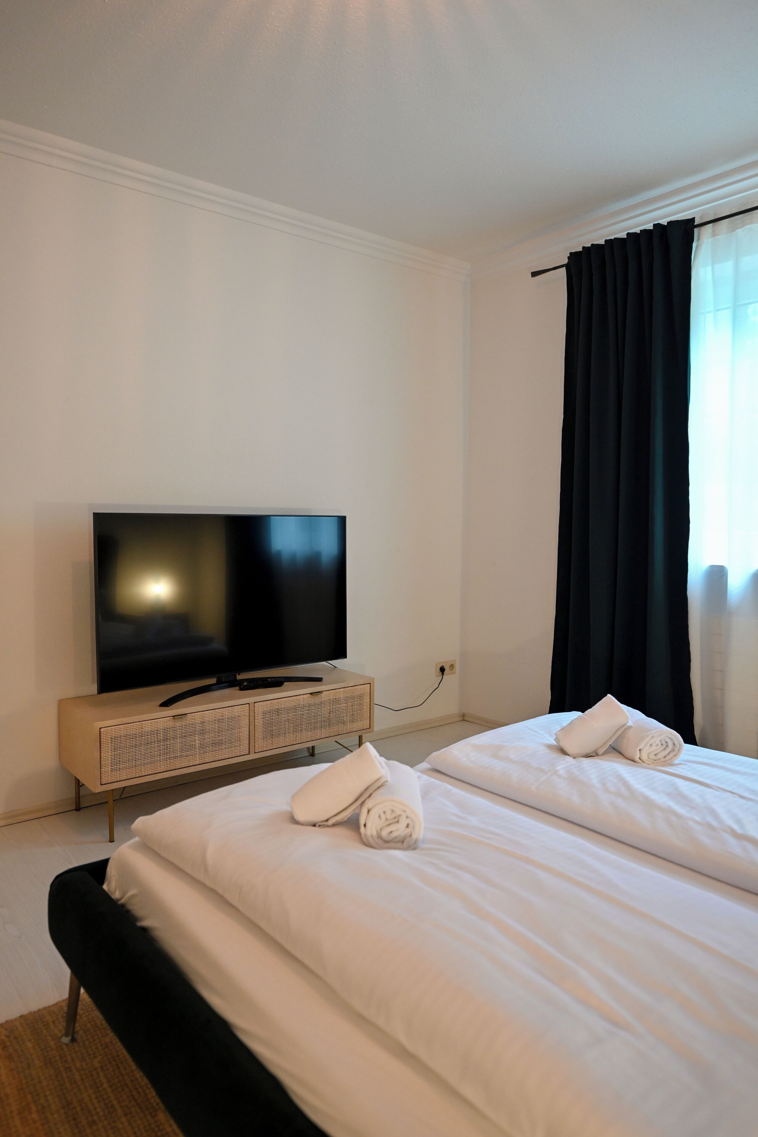 A hotel room with a flat-screen TV on a wooden stand, two beds with white linens, and a large window with black curtains.