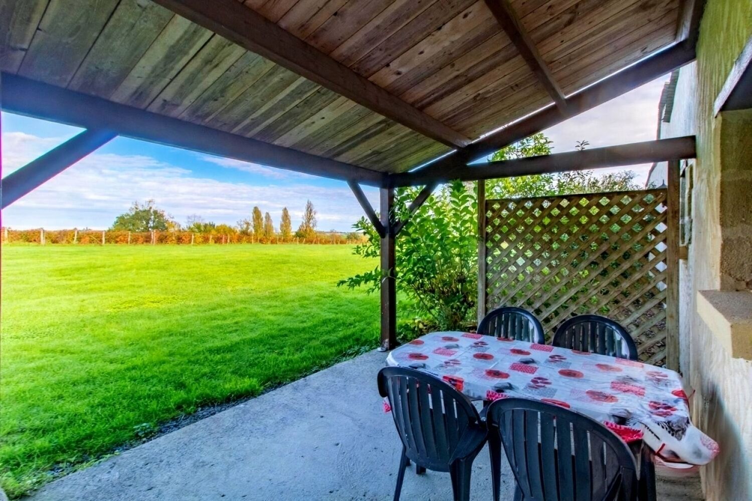 A covered outdoor area with a table and chairs, a view of a green field, and a wooden structure.