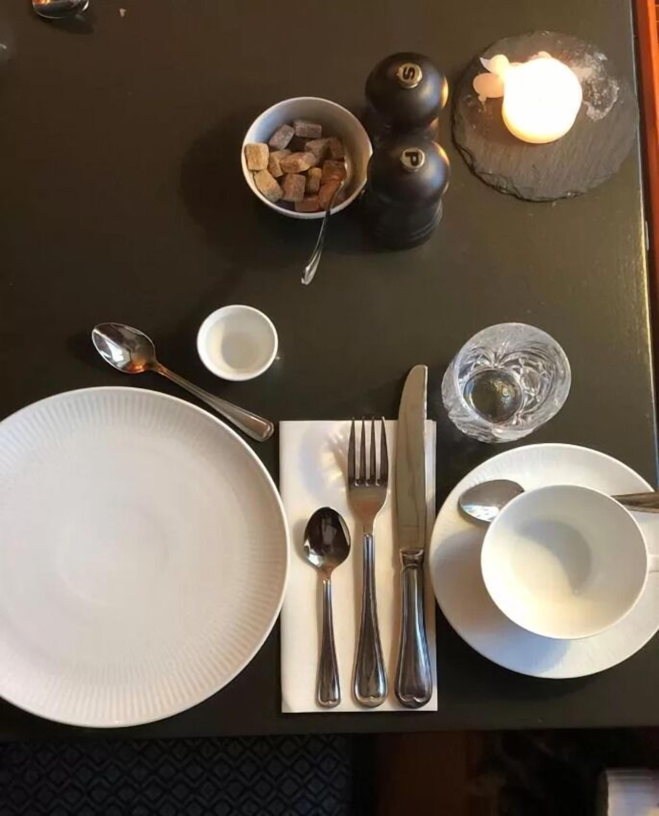 Une table à manger bien dressée, avec une assiette blanche, des couverts, un bol de morceaux de sucre, une bougie et un verre d’eau.