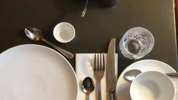Une table à manger bien dressée, avec une assiette blanche, des couverts, un bol de morceaux de sucre, une bougie et un verre d’eau.