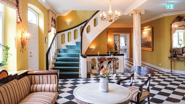A grand lobby with a checkered floor, a staircase, a sofa, a table with a vase, and a chandelier. - Hotel Baltic (Sydals)