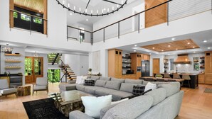 A spacious living area with a large grey sectional sofa, wooden flooring, and a modern kitchen with wooden cabinets and stainless steel appliances.