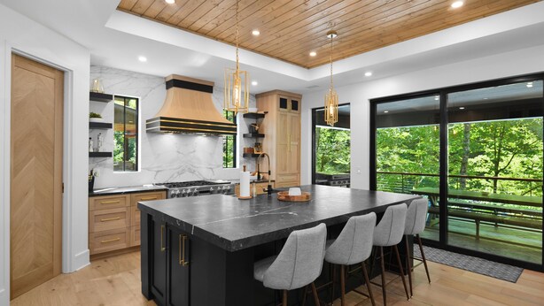 A modern kitchen with a large island, wooden ceiling, and large windows offering a view of the outdoors.