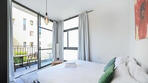 Et soveværelse med en stor seng, en balkon med bord og stole, og udsigt til bygninger.