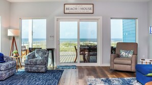 A spacious living room with a large sliding glass door leading to a deck with a view of the beach. There are two patterned armchairs and a blue sofa. - Modern Coastal Design ~ Sea La Vie (Mexico Beach)