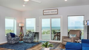 A spacious living room with a large window overlooking the beach, a ceiling fan, and a sign reading "Beach House." - Modern Coastal Design ~ Sea La Vie (Mexico Beach)