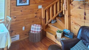 A wooden cabin interior with a staircase, framed pictures on the walls, a table with a chair, and a leather sofa.