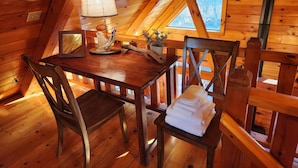 A cozy wooden cabin with a dining table, chairs, a lamp, and a view through a window.