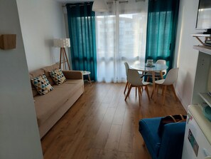 A living room with a sofa, a dining table with chairs, a lamp, and a TV. - Le Studiodé à Proximité Immédiate de la Plage et de la Thalasso (Bénodet)