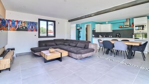 A modern living room with a sectional sofa, a coffee table, and a TV. Adjacent is a kitchen with a dining area, a microwave, and a sink. - Superbe Villa 6 Couchages + un lit Bébé, Jacuzzi, Piscine, Salle de Sport, Calme (Bouillargues)