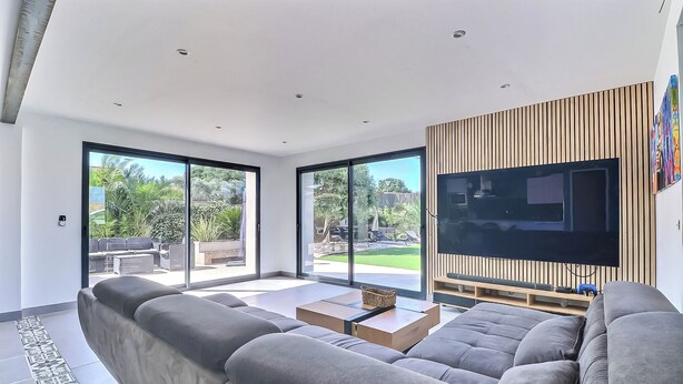 A modern living room with a large sofa, a coffee table, and a TV mounted on a wooden panel wall. - Superbe Villa 6 Couchages + un lit Bébé, Jacuzzi, Piscine, Salle de Sport, Calme (Bouillargues)