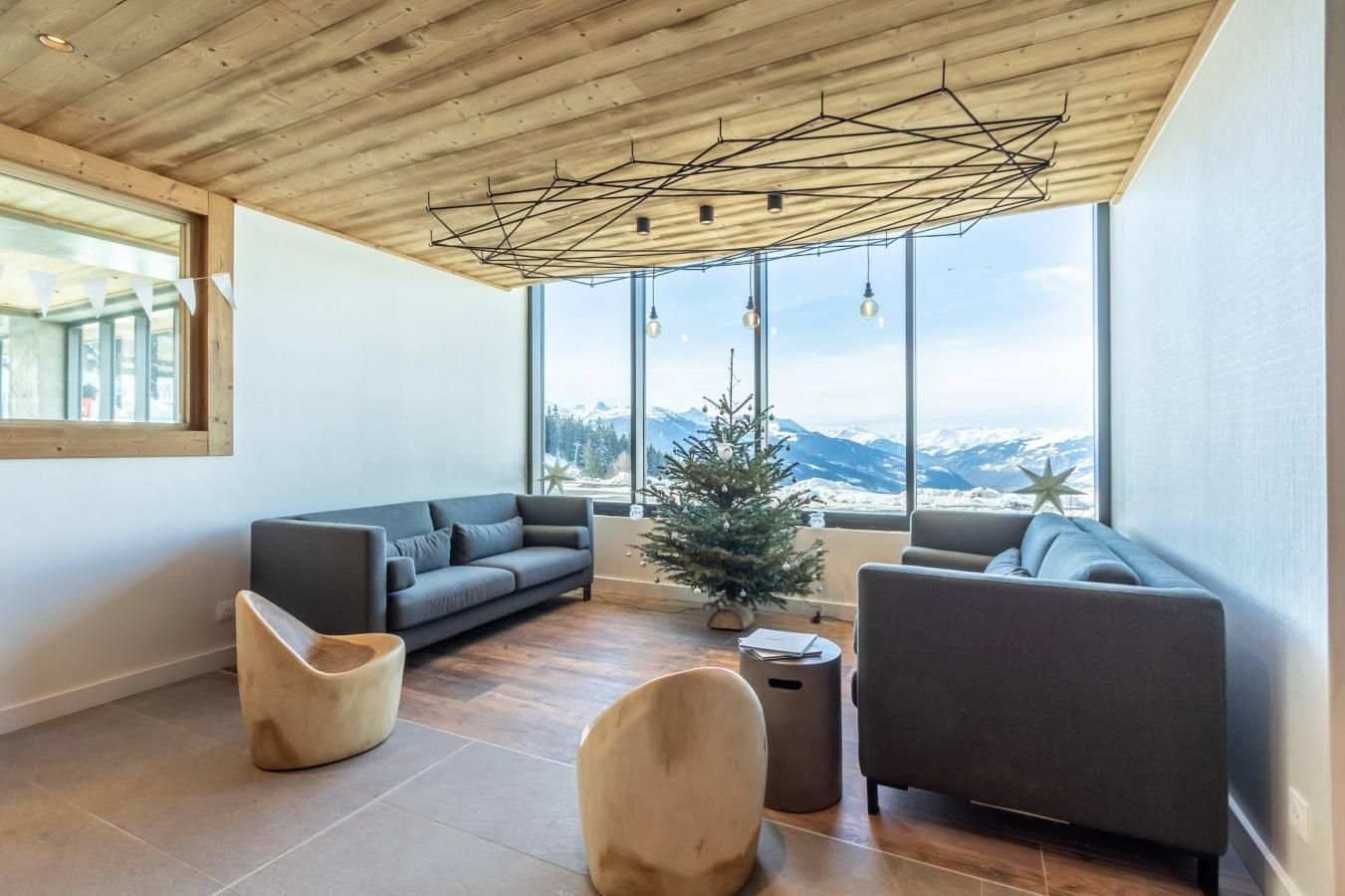 Una sala de estar moderna con techo de madera, ventanas grandes con vistas a la montaña y un pequeño árbol de Navidad.