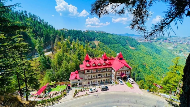 A hotel with a red roof, surrounded by lush green hills and a clear sky. - Hotel shimla hills international (Theog)