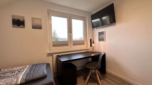 A bedroom with a bed, a desk, a chair, a window, and a TV on the wall. - B&W Apartments - DG Wohnung (Morschen)