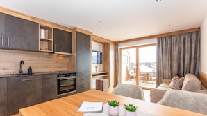 Een moderne keuken met donkere kastjes, een houten aanrechtblad en een inbouwoven. Er staat een eettafel met een placemat en twee kleine planten in potten. Een zithoek met een bank en uitzicht op een balkon is zichtbaar.