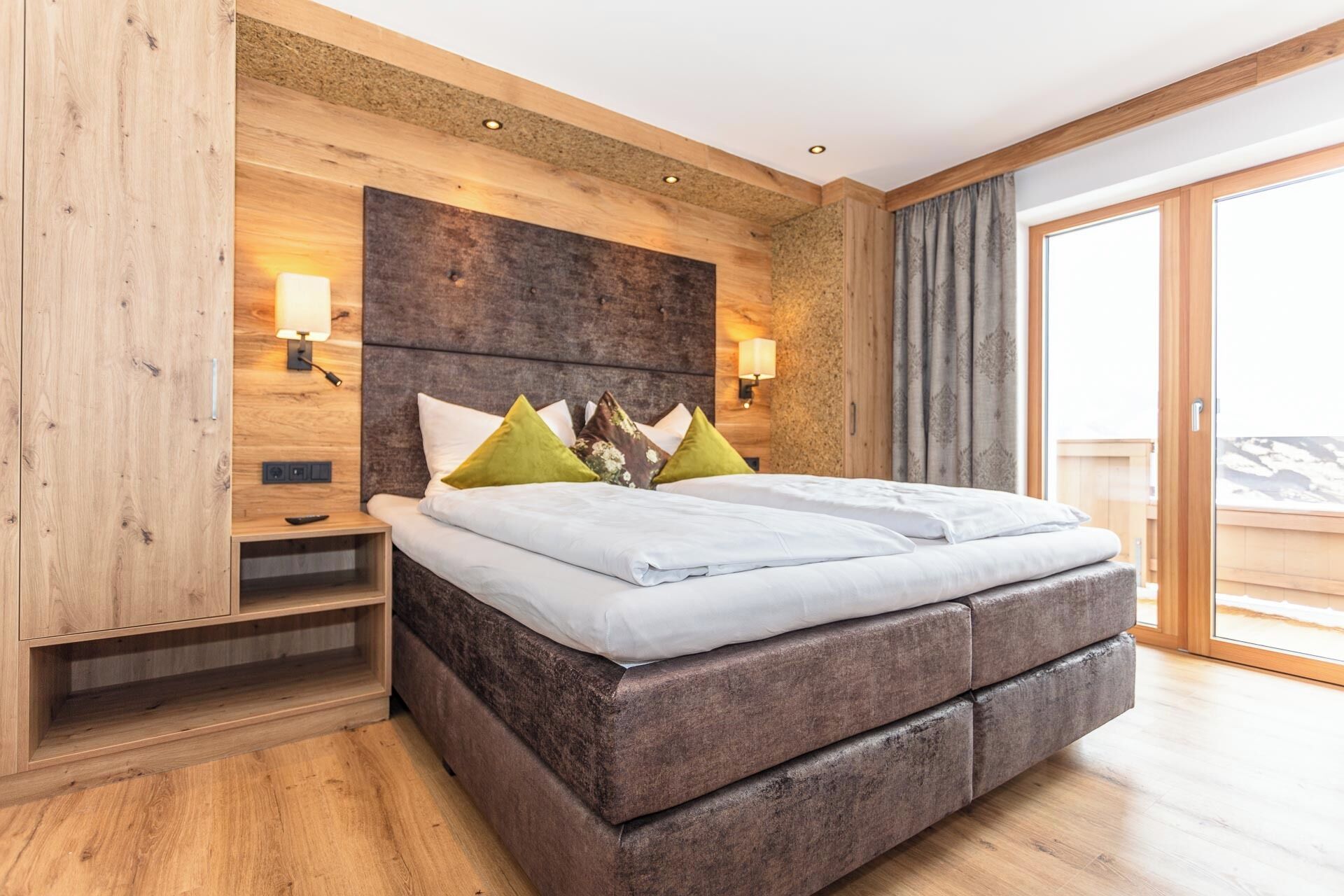 A modern bedroom with a wooden headboard, a large bed with white bedding, two bedside lamps, and a view of a balcony.