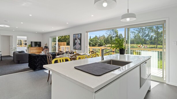 A modern kitchen with a large island, a dining area with yellow chairs, and a view of a garden through large windows. - The Whitehouse - Lepperton Holiday Home (New Plymouth)