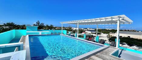 A rooftop infinity pool with a wooden deck, lounge chairs, and a pergola.