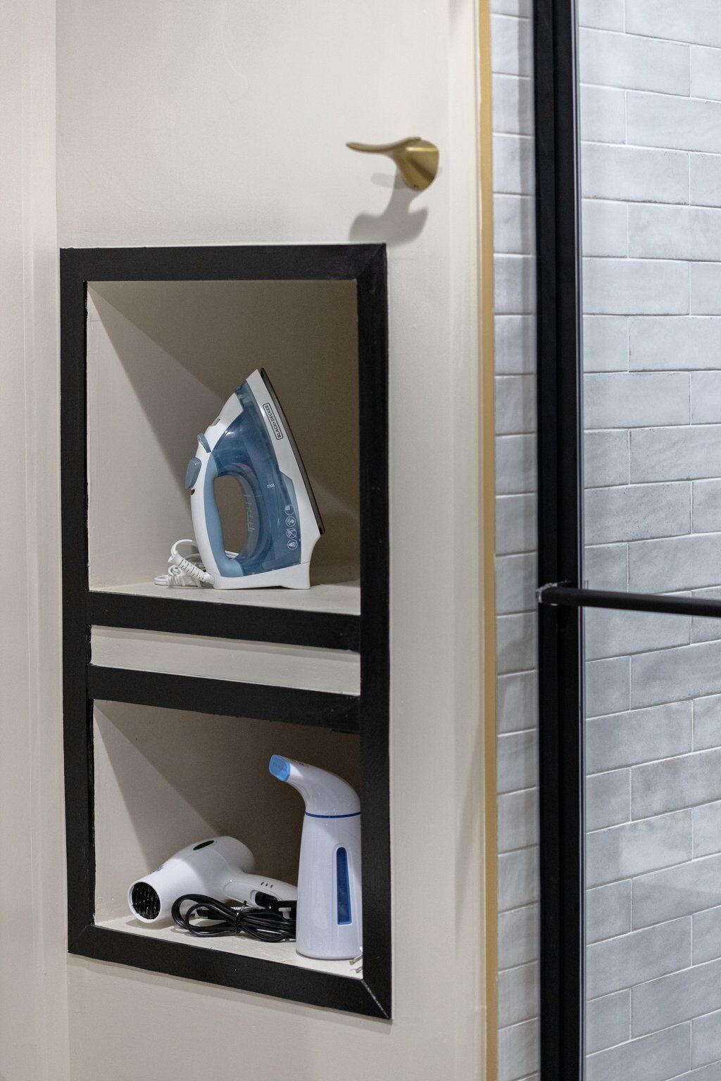 A door with a black frame and a white door handle. Inside the frame, there is a shelf with an iron, a hairdryer, and a spray bottle.