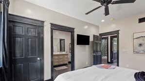 Un dormitorio con una cama grande, un ventilador de techo, un televisor de pantalla plana y una puerta que da a un baño.