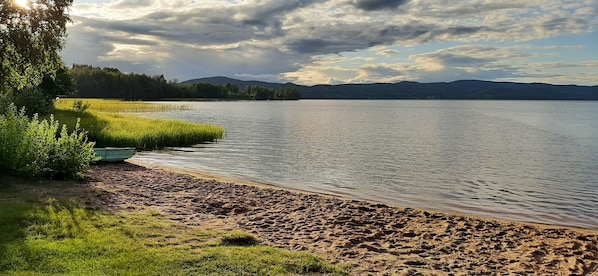 Blick auf einen See mit Sandstrand, Wiesen und einer Baumreihe.