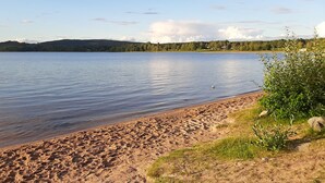 A sandy beach with calm water, a clear sky, and distant hills. - Vacation home with lake view, log cabin sauna and jacuzzi (Lakene)