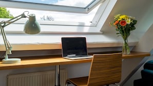 Une lucarne au-dessus d’un bureau en bois, sur lequel se trouvent un ordinateur portable, une lampe de bureau et un vase de fleurs.