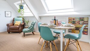 Un espace repas doté d’un plafond haut et de lucarnes, comprenant une table ronde entourée de chaises, une plante suspendue et un mur orné d’étiquettes anciennes de produits alimentaires.