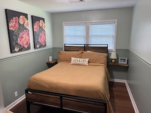 A bedroom with a bed, a nightstand, a window with blinds, and two floral paintings on the wall. - 3 Bed, 1 Bath Adorable Home – Less than 10 minutes from Bryant-Denny Stadium! (Northport)