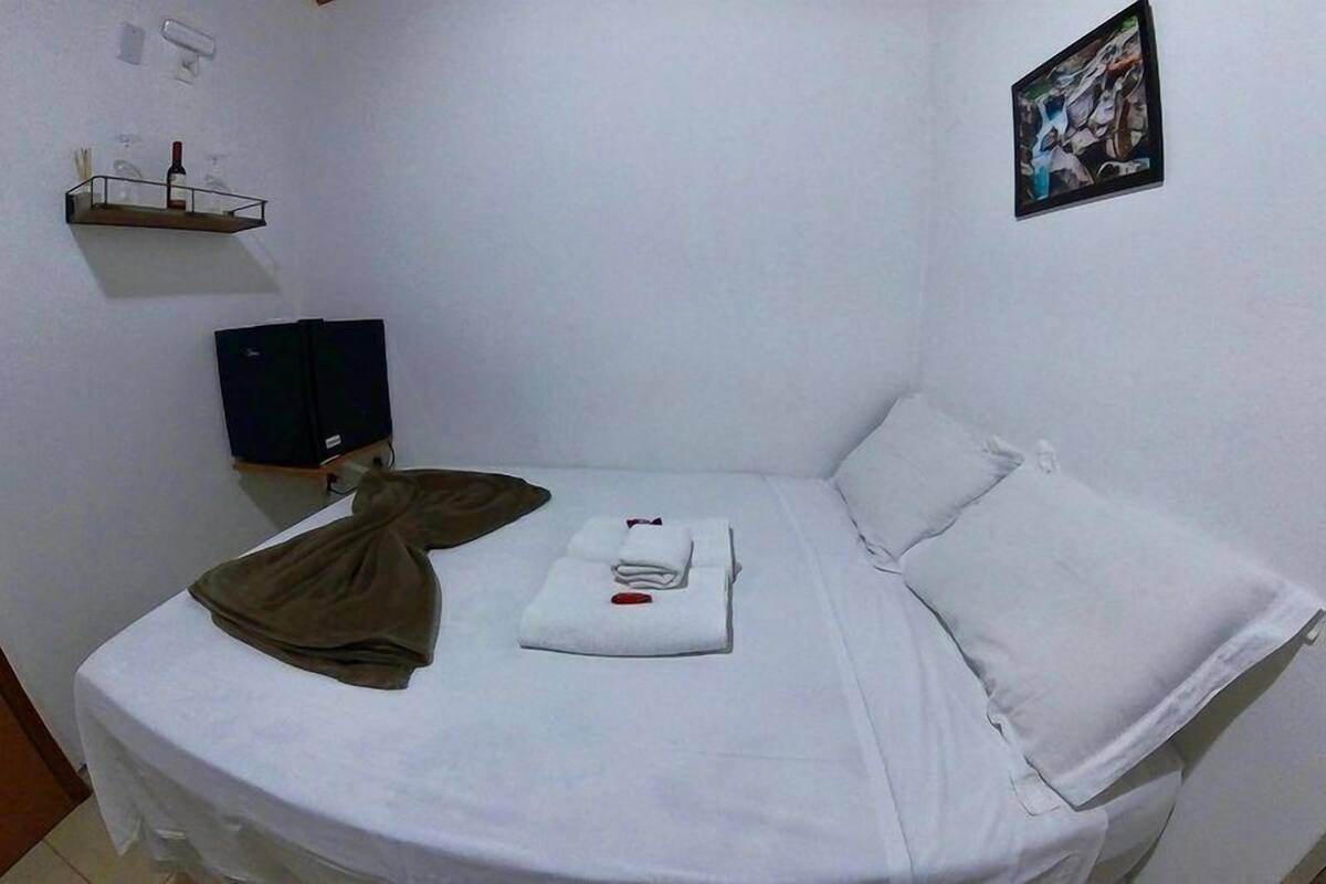 Un petit lit bien fait, avec des draps blancs, une veste marron et une serviette pliée posée sur le lit. Sur le mur, on aperçoit une étagère avec une bouteille et des verres, et au-dessus du lit, un tableau encadré.